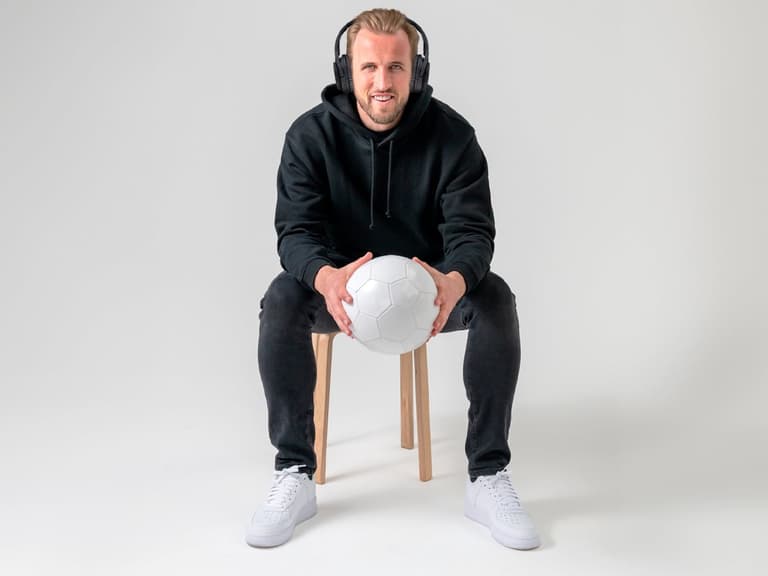 Harry Kane wearing headphones, sitting on a stool, holding a white soccer ball, in a studio setting.