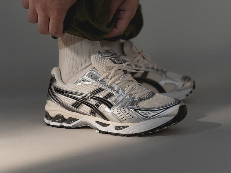 Asics Gel Kayano 14 sneakers in silver and black worn with white socks and green pants.