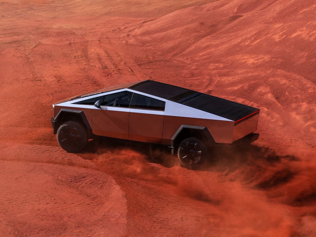 Tesla Cybertruck driving on red desert terrain with dust trailing behind, showcasing its angular stainless steel design.