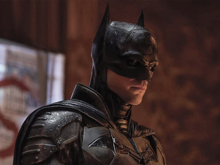Batman in dark suit stands in dimly lit room, showcasing detailed armour and iconic cowl.