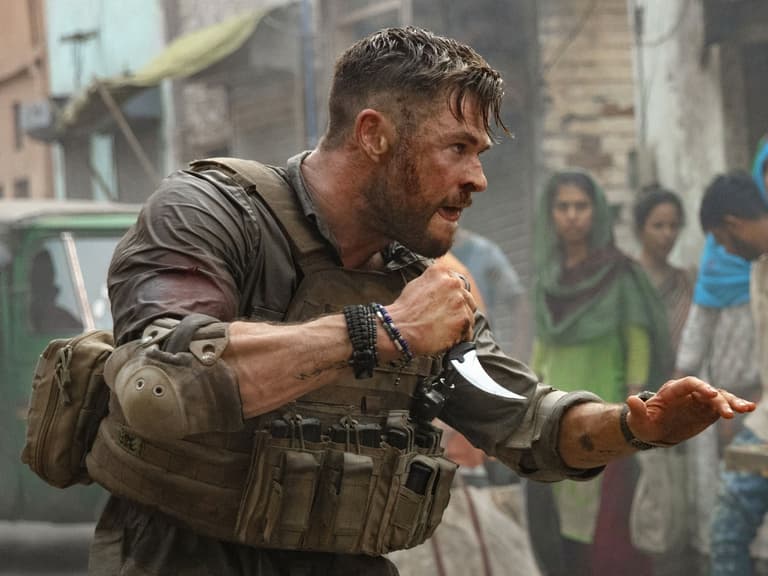 Chris Hemsworth in combat gear holding a knife, surrounded by onlookers in a gritty urban setting.