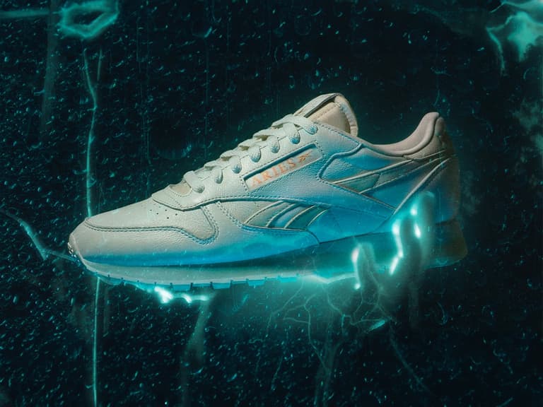 Aries Reebok sneaker in white leather with glowing blue accents and textured background.