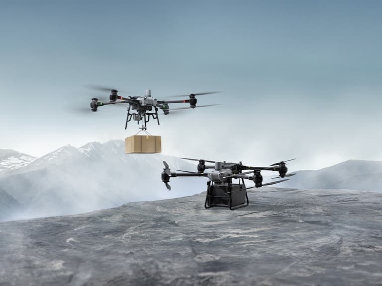DJI FlyCart 30 drones hover over a rocky landscape, one carrying a package, under a cloudy sky.