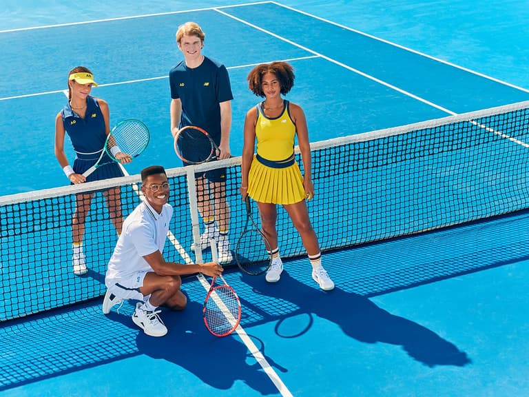 Tennis players in New Balance x AO 2024 apparel pose on a bright blue court, holding racquets and smiling.