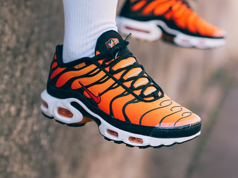Nike Air Max Plus TN in orange and black with white sole, worn outdoors against a textured wall.