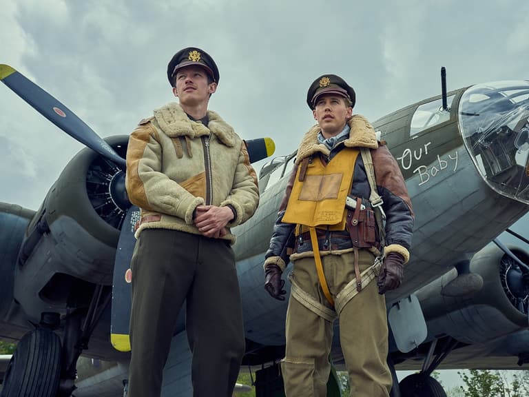Two men in vintage bomber jackets stand in front of a military aircraft with "Our Baby" painted on the nose.