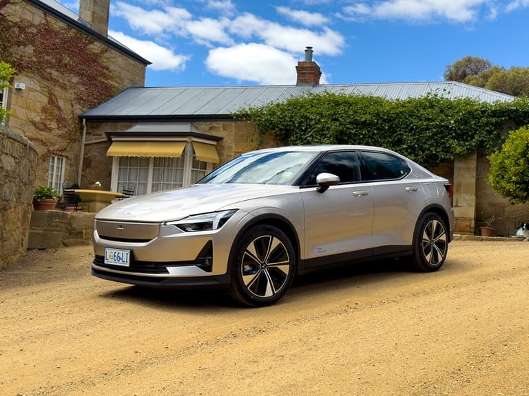 Polestar 2 in silver parked on a gravel driveway in front of a rustic stone house under a blue sky.