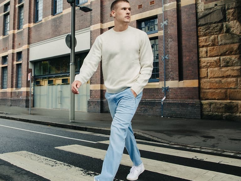 Man in cream Uniqlo sweater and light blue jeans crossing a city street, with a brick building in the background.