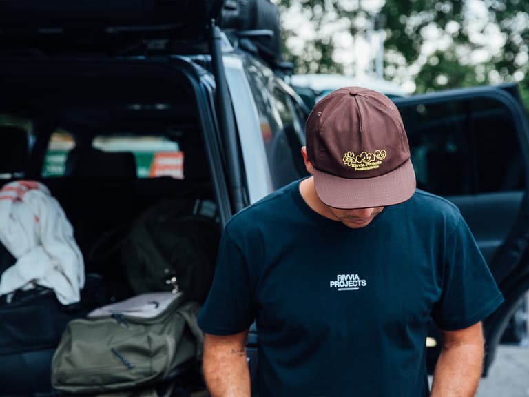 Man wearing Rivvia Projects cap and T-shirt stands near an open car boot, with trees in the background.