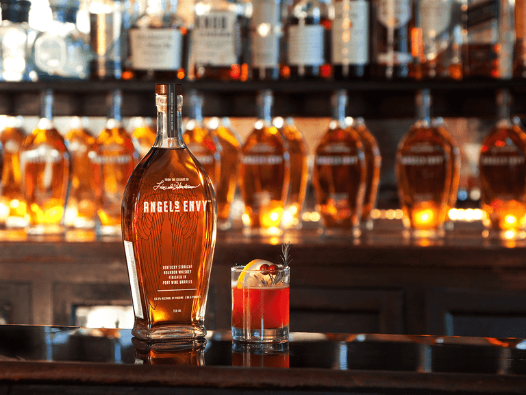 Angel's Envy bottle and cocktail on a bar with backlit whisky bottles.
