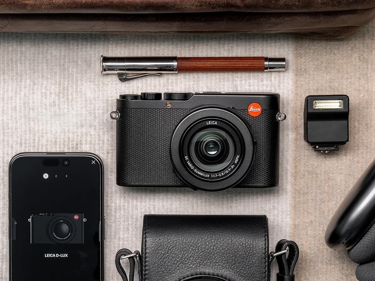 Leica D-Lux 8 camera in black, surrounded by a smartphone, pen, and camera accessories on a textured surface.