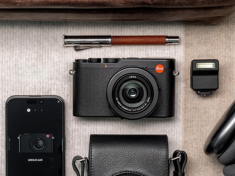 Leica D-Lux 8 camera in black, surrounded by a smartphone, pen, and camera accessories on a textured surface.