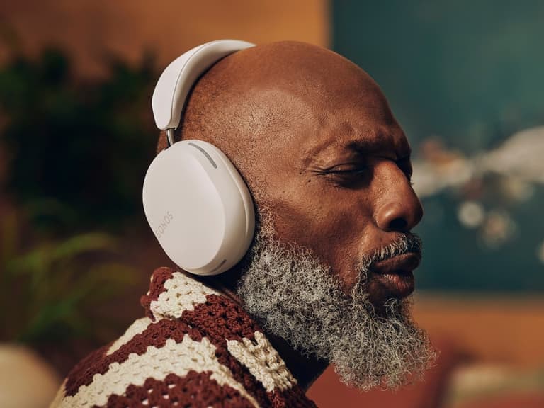 Man wearing Sonos Ace headphones, eyes closed, in a cosy room with warm lighting.