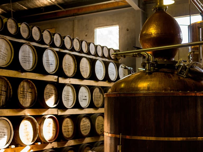 Sullivans Cove distillery interior with wooden barrels stacked against a wall and a copper still in warm lighting.