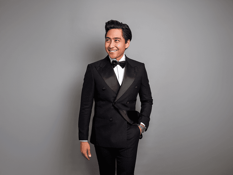 Theodore tailor wearing a black double-breasted suit and bow tie, smiling against a grey background.