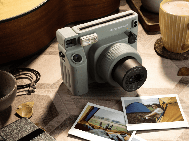 Fujifilm Instax Wide 400 camera in grey on a wooden table with instant photos, guitar, and coffee cup nearby.