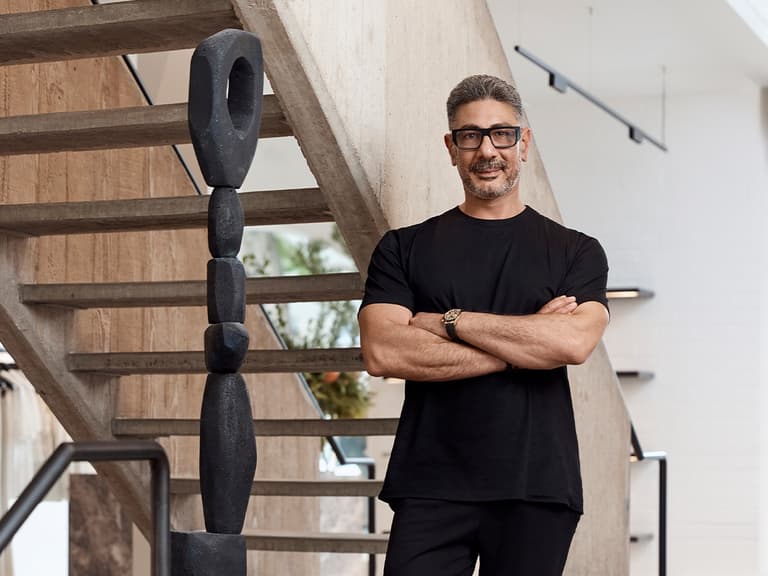 Joe Farage stands confidently in a modern interior, wearing a black t-shirt and glasses, with arms crossed.