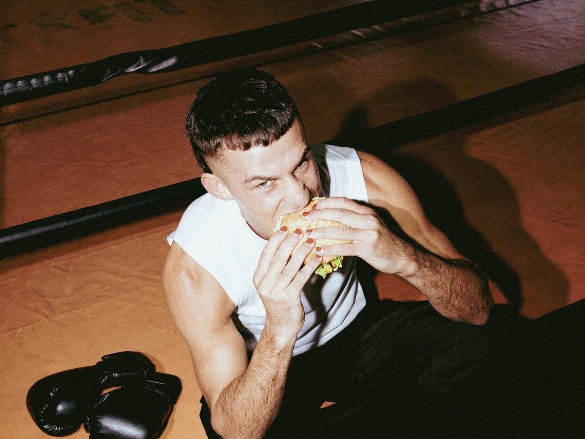 Harry Garside eating a sandwich while sitting on a boxing ring floor, with boxing gloves beside him.