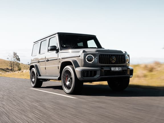2025 Mercedes-AMG G 63 in grey driving on a rural road with blurred motion, under clear skies.