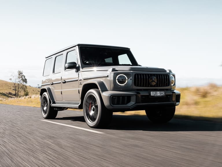 2025 Mercedes-AMG G 63 in grey driving on a rural road with blurred motion, under clear skies.