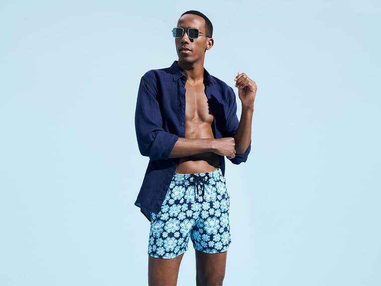 Man wearing blue floral swim shorts and navy shirt against a light blue background.