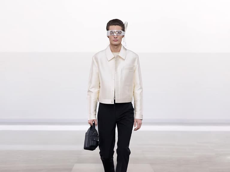 Model wearing Dior cream jacket and black trousers walks runway with a black handbag, minimalist backdrop.