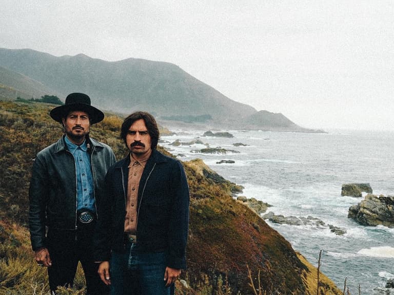 Hermanos Gutiérrez stand on a coastal cliff with rugged terrain and ocean waves in the background.