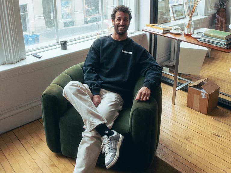 Daniel Ricciardo smiling in a green armchair, wearing a black Enchanté sweatshirt and white pants in a bright room.