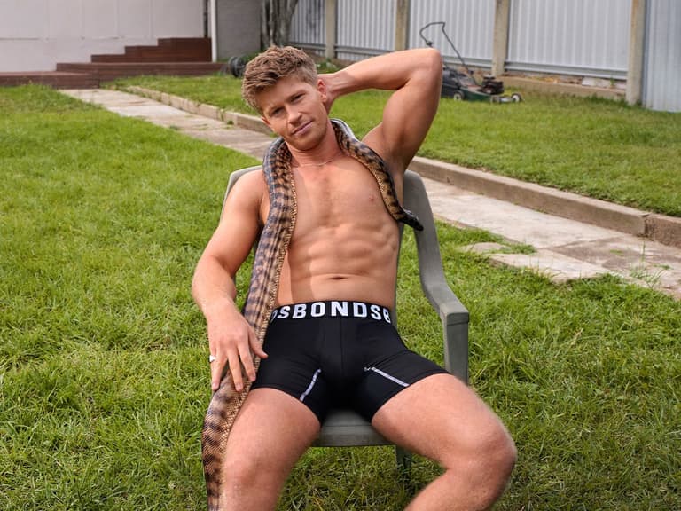 Robert Irwin sits on a chair outdoors wearing black Bonds underwear with a snake draped around his neck.