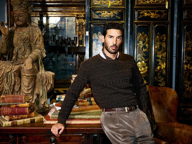 Man in brown patterned sweater leans on antique table surrounded by vintage books and ornate Asian decor.