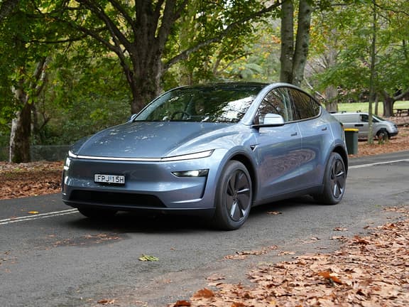 Tesla Model Y in blue driving on a tree-lined road with fallen leaves, showcasing its sleek design.