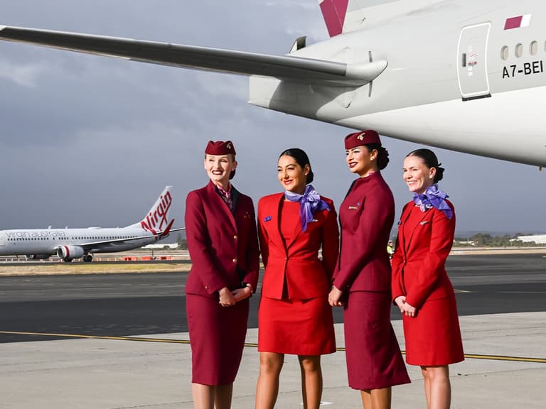 Virgin Australia flight attendants in red uniforms stand smiling on tarmac near aircraft tail.