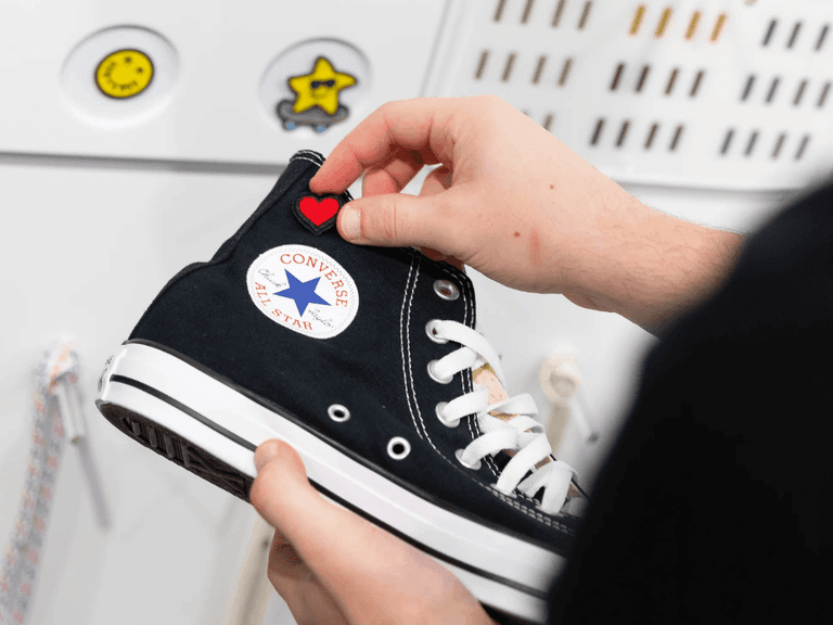 Person customising black Converse Chuck Taylors with red heart patch, white wall background.