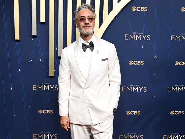 Man in a double-breasted white suit with a black bow tie, standing on a blue Emmy Awards backdrop.
