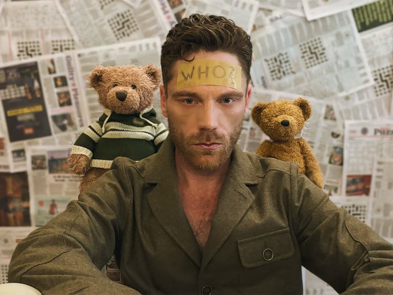 Scott Rose-Marsh wearing a brown jacket with two teddy bears on his shoulders, sitting in front of a newspaper-covered wall.
