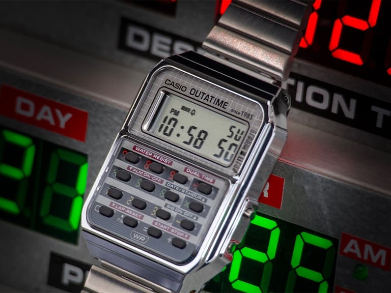 Casio CA500WEBF-1A watch with silver metal band and digital display, set against a futuristic backdrop with red and green lights.