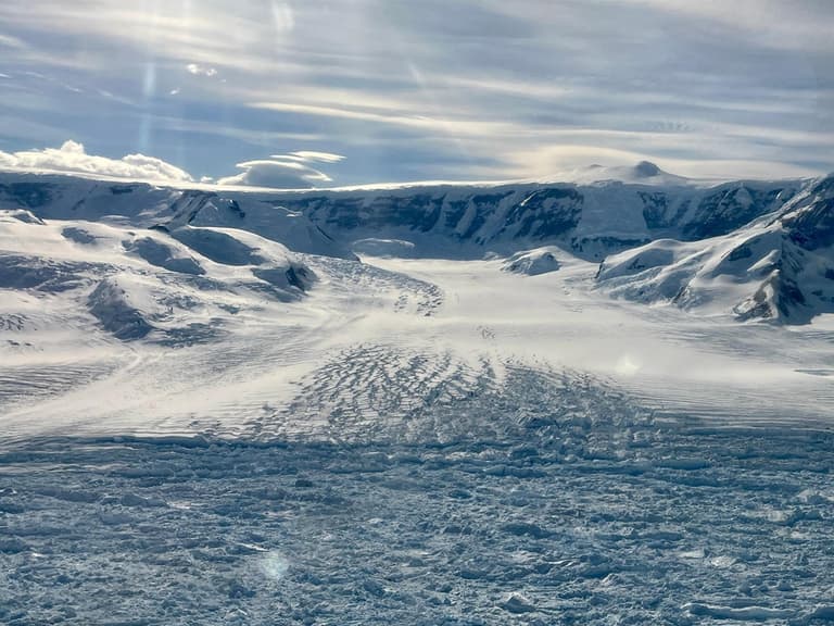 Antarctic glacier with visible crevasses under a bright, cloudy sky, highlighting retreat and ice loss.