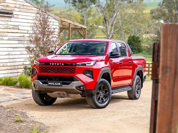 2026 Toyota HiLux Rogue in red, front three-quarter view, parked on a rural dirt driveway with a wooden building nearby.