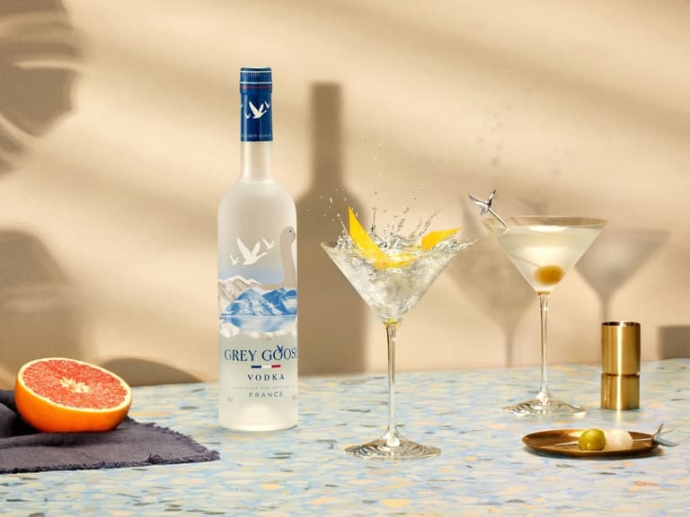 Grey Goose vodka bottle with two martinis, garnished with lemon twist and olive, on a terrazzo surface.