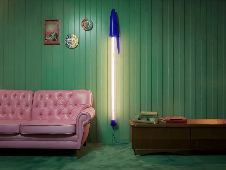 Seletti Bic Pen Lamp in blue and white, mounted on a green panelled wall beside a pink sofa and wooden coffee table.