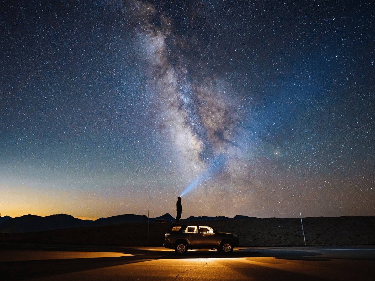 Person standing on a vehicle shines flashlight into starry night sky with Milky Way visible above.