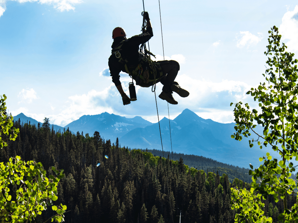 A person rappelling with mountains in the background, surrounded by lush green trees under a bright sky.