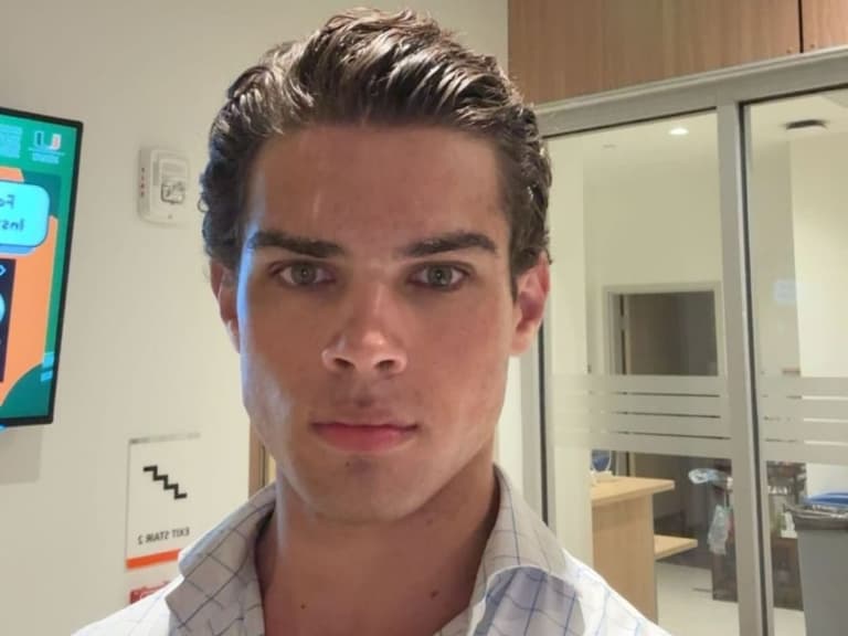 Man with slicked-back hair and a serious expression stands indoors, wearing a checkered shirt, with office decor in the background.