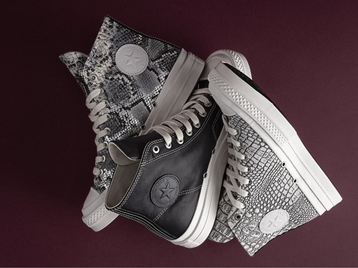 Converse First String sneakers in black leather and grey snakeskin on a dark maroon background.