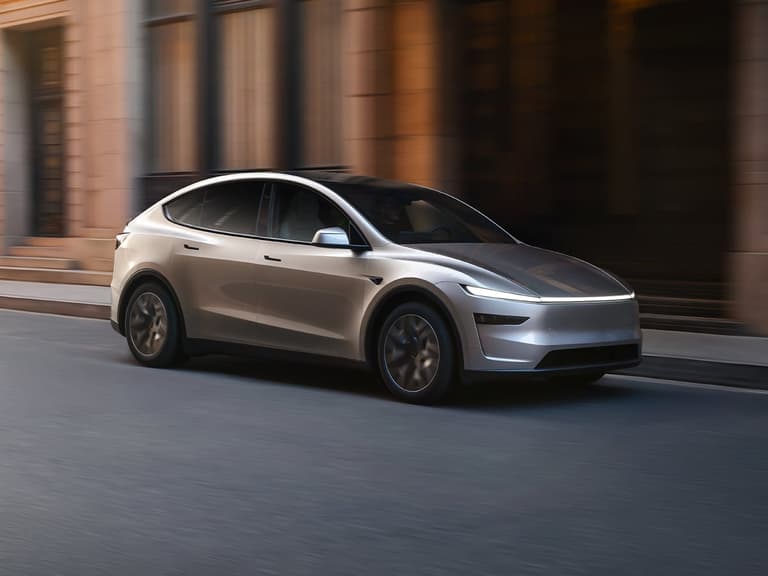 Tesla Model Y L in silver driving on a city street with blurred background and warm lighting.