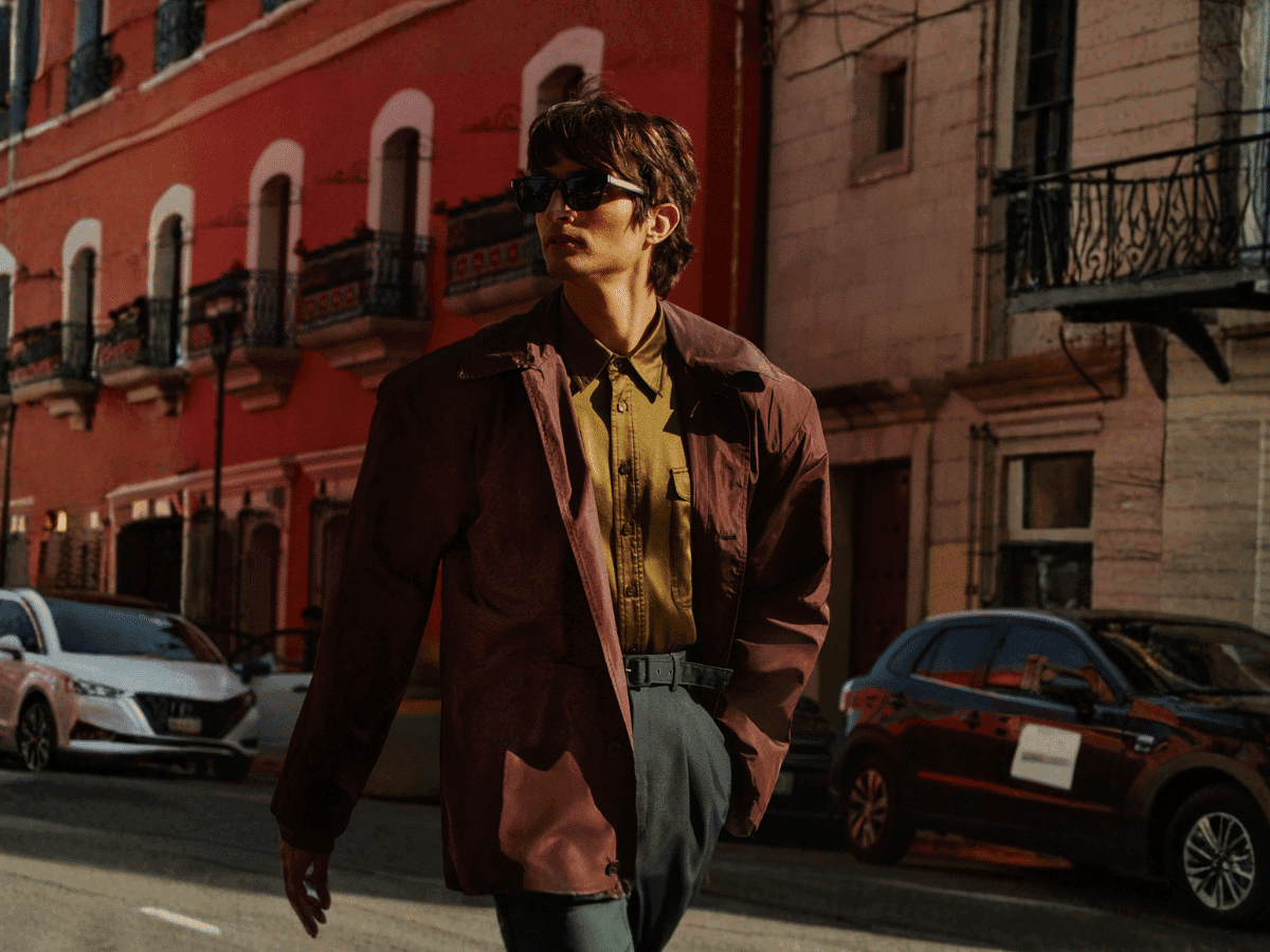 Man in brown jacket and sunglasses walking on a city street with red and beige buildings in the background.