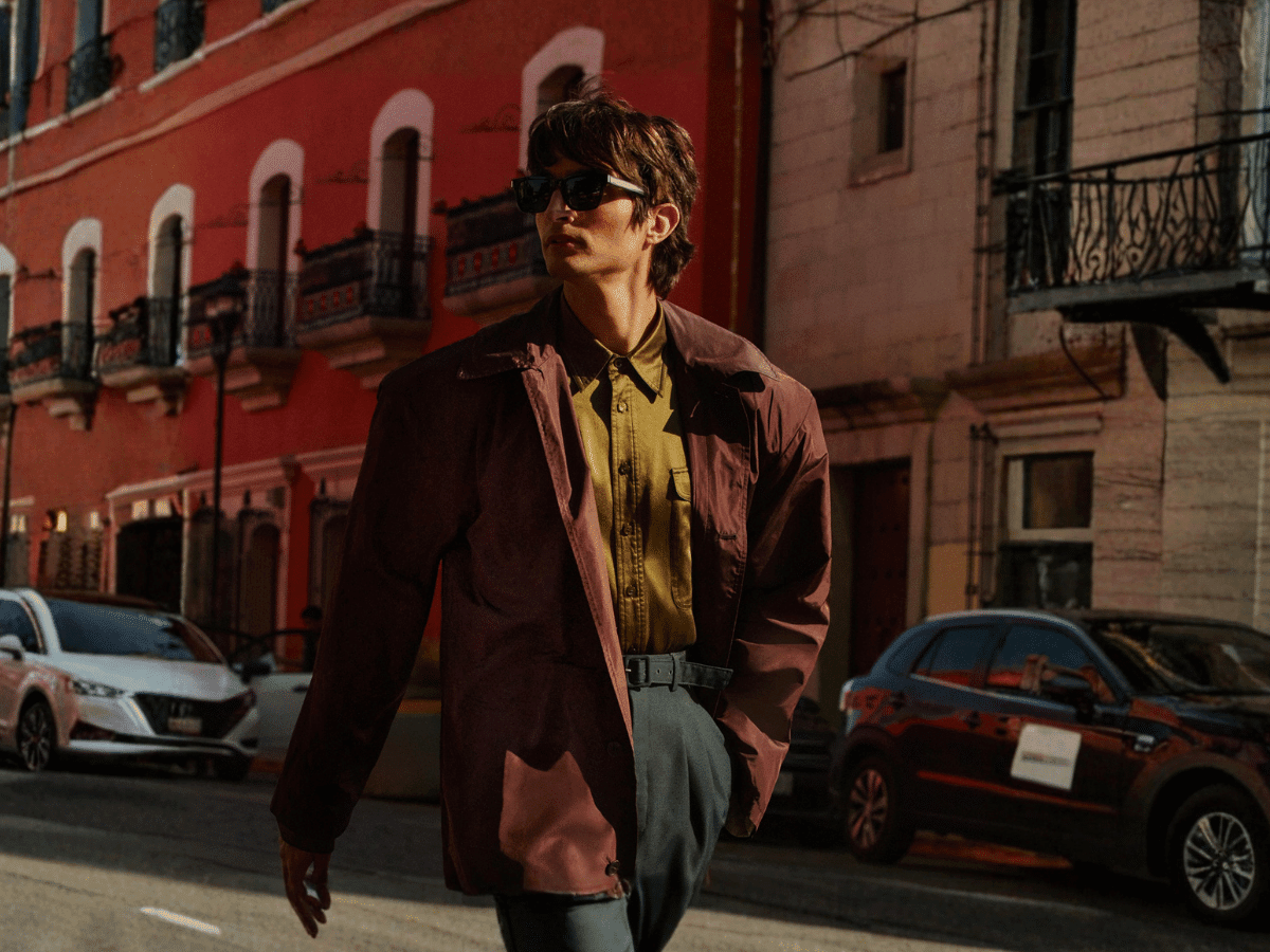 Man in brown jacket and sunglasses walking on a city street with red and beige buildings in the background.