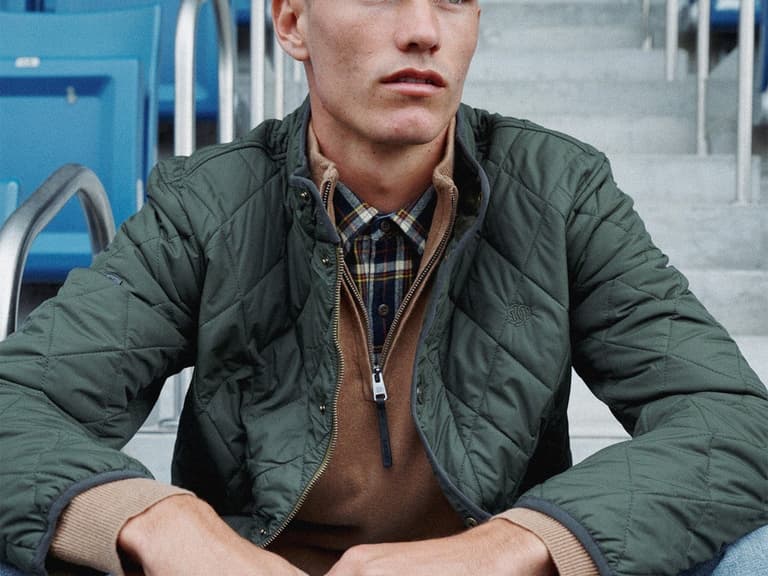 Man wearing a quilted olive Superdry jacket over a hoodie, seated in stadium stands