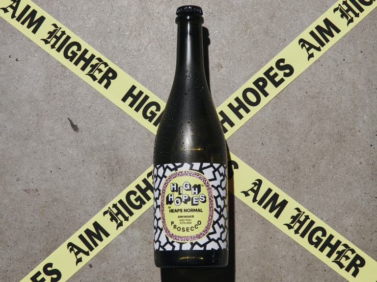 Bottle of High Hopes prosecco centred in a yellow Aim Higher / High Hopes tape X on concrete