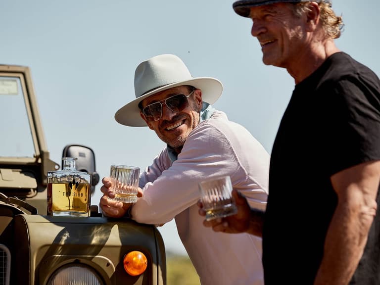 Two men sharing glasses of The Bird whiskey with the bottle on a vehicle bonnet